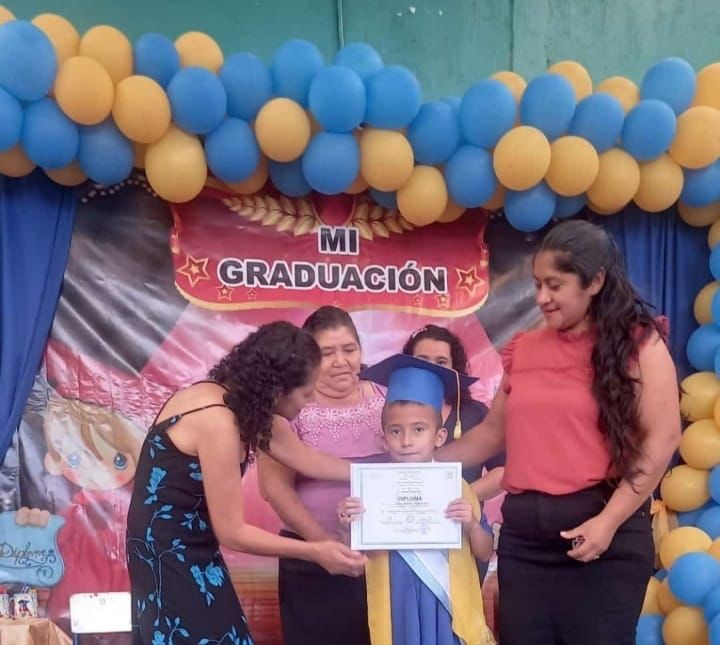 Graduation ceremony with a crowd of people around a young boy. They are handing him a graduation certificate.